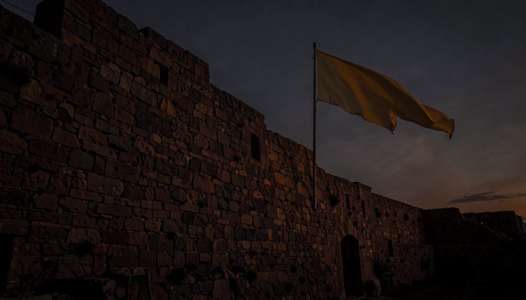 Stone fortress wall at dusk with a large yellow flag waving against a darkening sky, dramatic silhouette mood