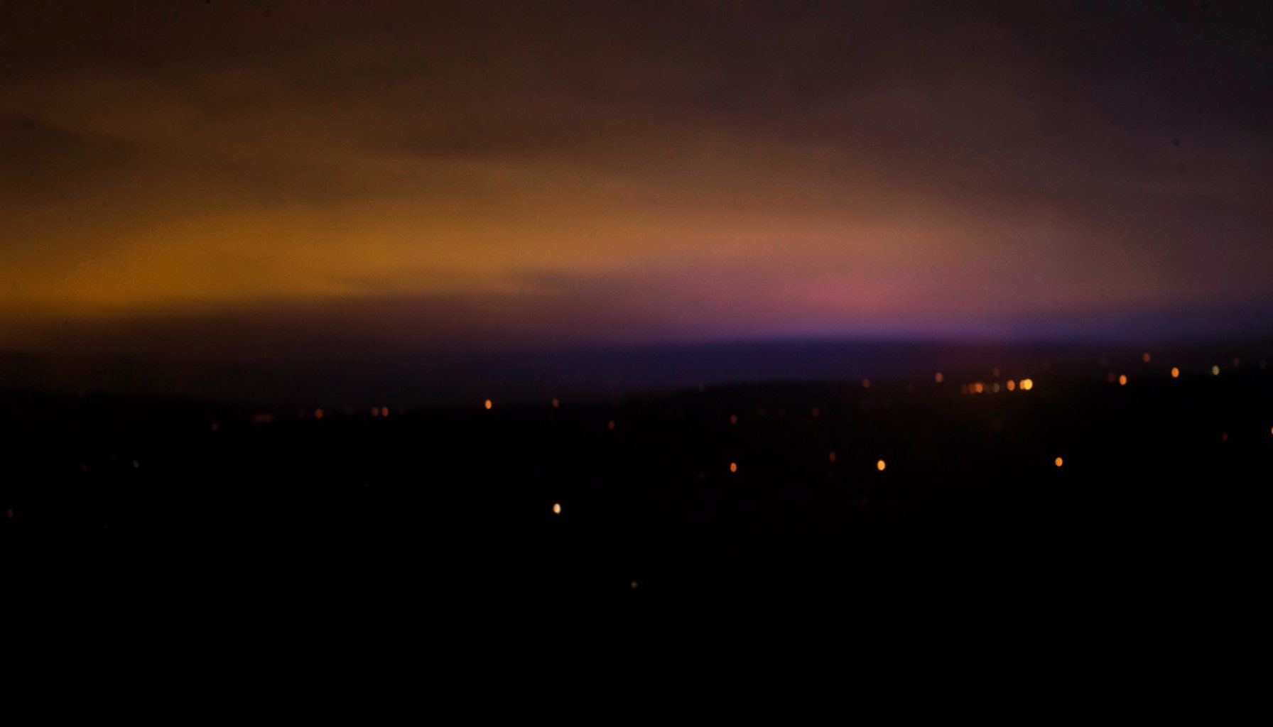 Blurry night skyline view with distant lights and colorful sunset glow in orange, purple, and blue tones, moody atmosphere