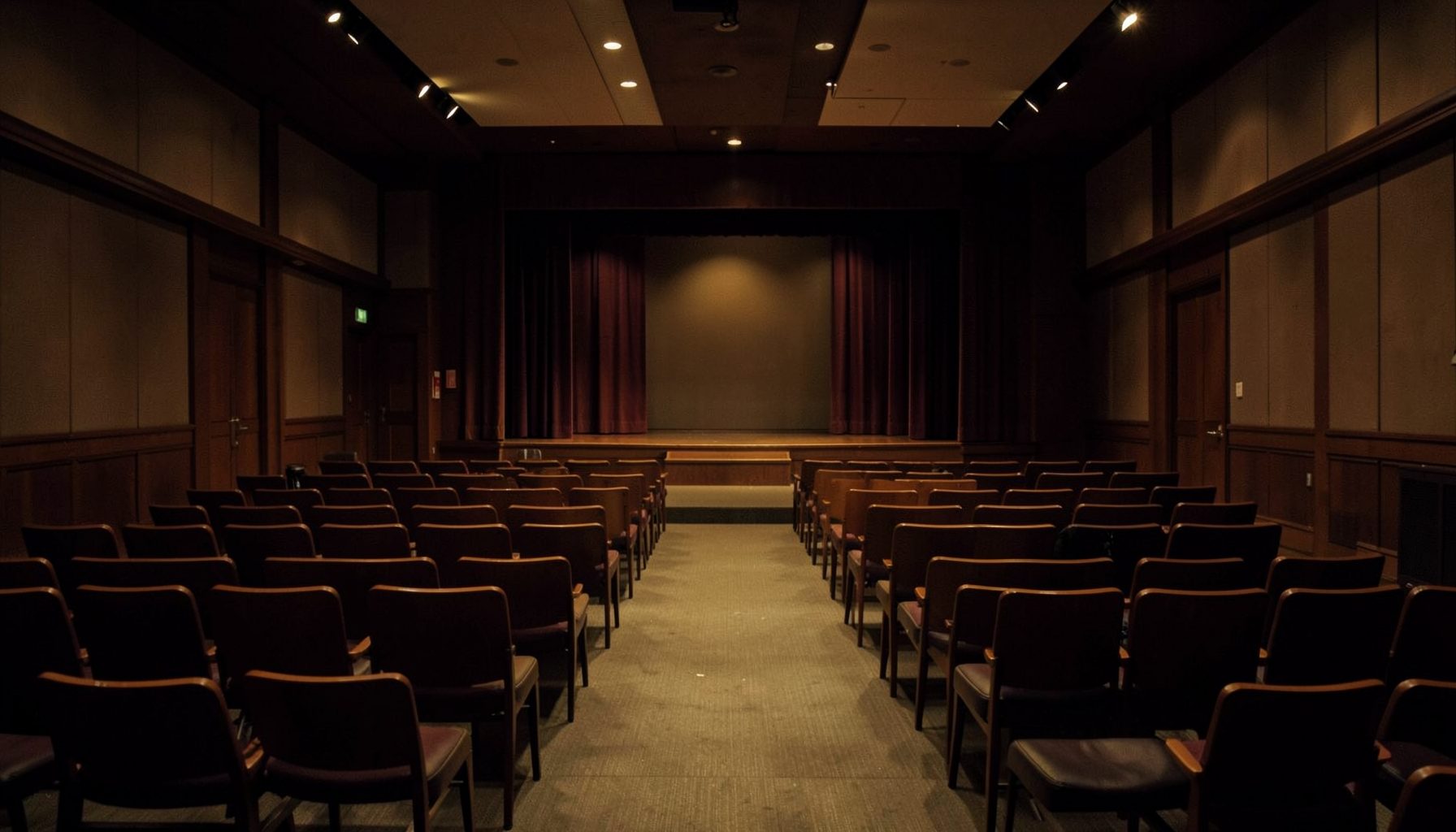 Empty theater or auditorium with rows of chairs facing a curtained stage, dim warm lighting, quiet mood