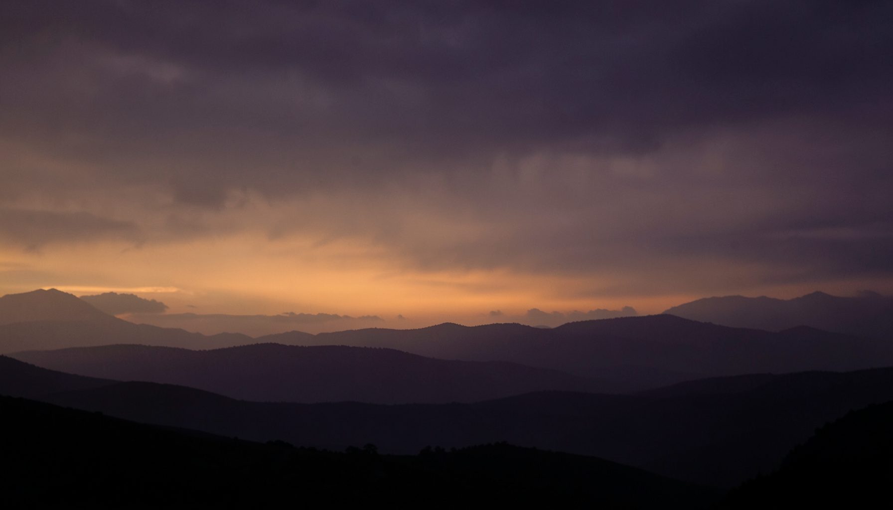 Dramatic cloudy sky over mountain silhouettes at dusk, purple and orange tones, moody atmosphere
